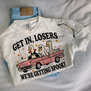 K&ouml;nnte beinhalten: Cremefarbenes T-Shirt mit dem Text "GET IN, LOSERS WE'RE GETTING SPOOKY" und einer Grafik von drei Enten in einem rosa Auto, verkleidet als Vampir, Geist und Mumie. Das Shirt wird mit einer blauen Jeans kombiniert.