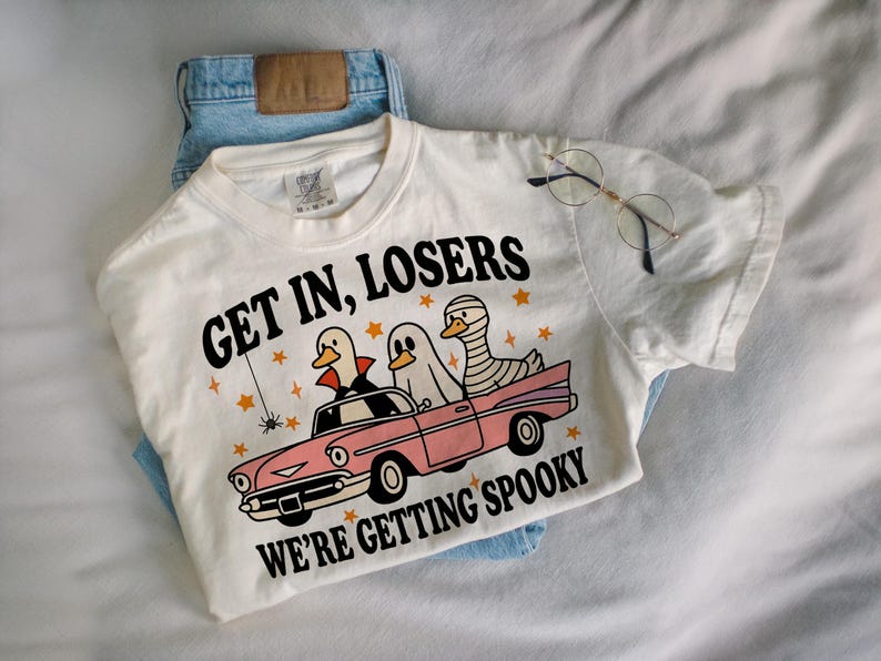 K&ouml;nnte beinhalten: Cremefarbenes T-Shirt mit dem Text "GET IN, LOSERS WE'RE GETTING SPOOKY" und einer Grafik von drei Enten in einem rosa Auto, verkleidet als Vampir, Geist und Mumie. Das Shirt wird mit einer blauen Jeans kombiniert.