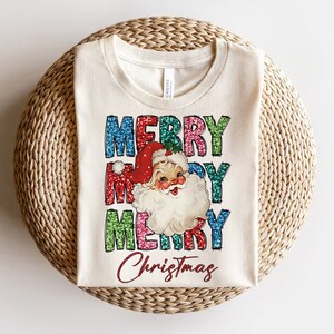 Op de afbeelding: Een wit T-shirt met een vintage-stijl illustratie van de kerstman en de woorden "Merry Christmas" in kleurrijke, glinsterende letters.