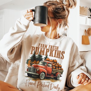 Op de afbeelding: Een wit sweatshirt met een vintage truck-ontwerp. De truck is rood en heeft een laadbak vol pompoenen. De tekst op het sweatshirt luidt "Farm Fresh Pumpkins Hand Picked Daily".