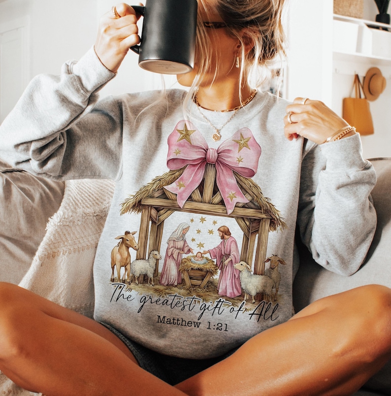 May include: A gray sweatshirt with a pink bow and a nativity scene design. The text "The greatest gift of all" and "Matthew 1:21" are printed below the scene.