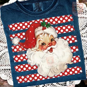 May include: A dark blue t-shirt with a vintage-style graphic of Santa Claus wearing a red hat with a white pom-pom and holly leaves. The background of the graphic features horizontal red stripes with white polka dots across the chest area.