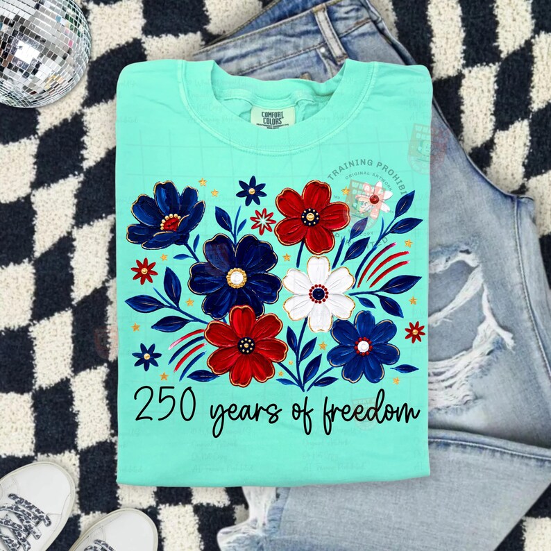 May include: A light turquoise t-shirt with a floral design in red, white, and blue. The design includes the text "250 years of freedom". The shirt is folded on top of a pair of blue jeans.