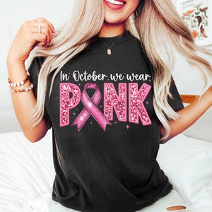 May include: Black t-shirt with the text "In October we wear PINK" in pink glitter letters with a pink ribbon in the center.