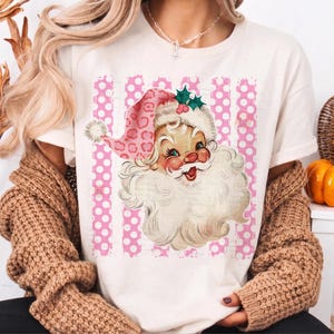 Pode incluir: Uma t-shirt de cor creme com um gr&aacute;fico de um Pai Natal de estilo vintage, usando um gorro cor-de-rosa com padr&atilde;o de leopardo, um pompom branco e folhas de azevinho verdes. O fundo do gr&aacute;fico consiste em riscas verticais cor-de-rosa e brancas com bolinhas.