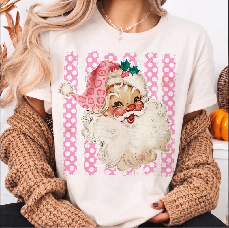 Pode incluir: Uma t-shirt de cor creme com um gr&aacute;fico de um Pai Natal de estilo vintage, usando um gorro cor-de-rosa com padr&atilde;o de leopardo, um pompom branco e folhas de azevinho verdes. O fundo do gr&aacute;fico consiste em riscas verticais cor-de-rosa e brancas com bolinhas.