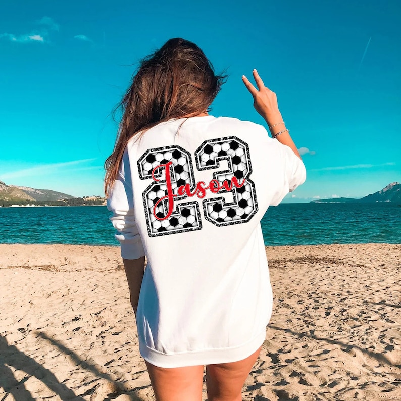 May include: White sweatshirt with a black and white soccer ball design featuring the number 23 and the name "Jason" in red.