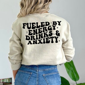May include: A beige sweatshirt with black text that reads "Fueled by Energy Drinks & Anxiety" in a retro font.