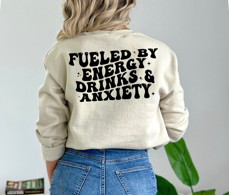 May include: A beige sweatshirt with black text that reads "Fueled by Energy Drinks & Anxiety" in a retro font.