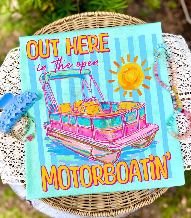 May include: A light blue t-shirt with a colorful illustration of a pontoon boat, a yellow sun, and the text "OUT HERE in the open MOTORBOATIN'". The shirt is decorated with vertical blue stripes.