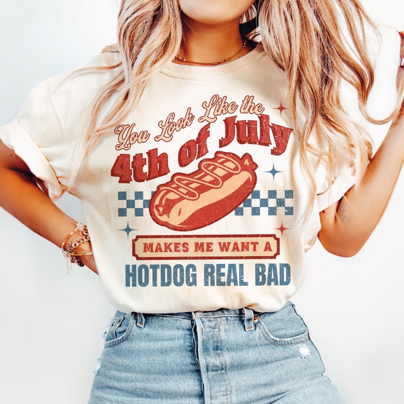 May include: A white t-shirt with a vintage-style graphic that says "You look like the 4th of July makes me want a hotdog real bad". The graphic features a red, white, and blue hotdog with mustard and ketchup.