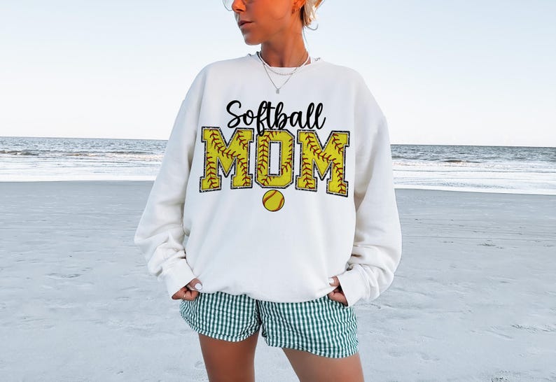 May include: White sweatshirt with the text "Softball MOM" in black and yellow. The letters are outlined in red and have a softball pattern.