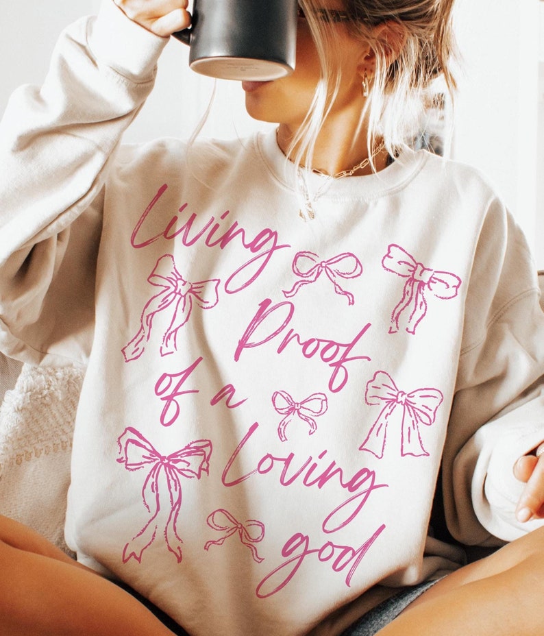 May include: A white sweatshirt with the text "Living Proof of a Loving God" in pink script. The text is surrounded by pink bows.