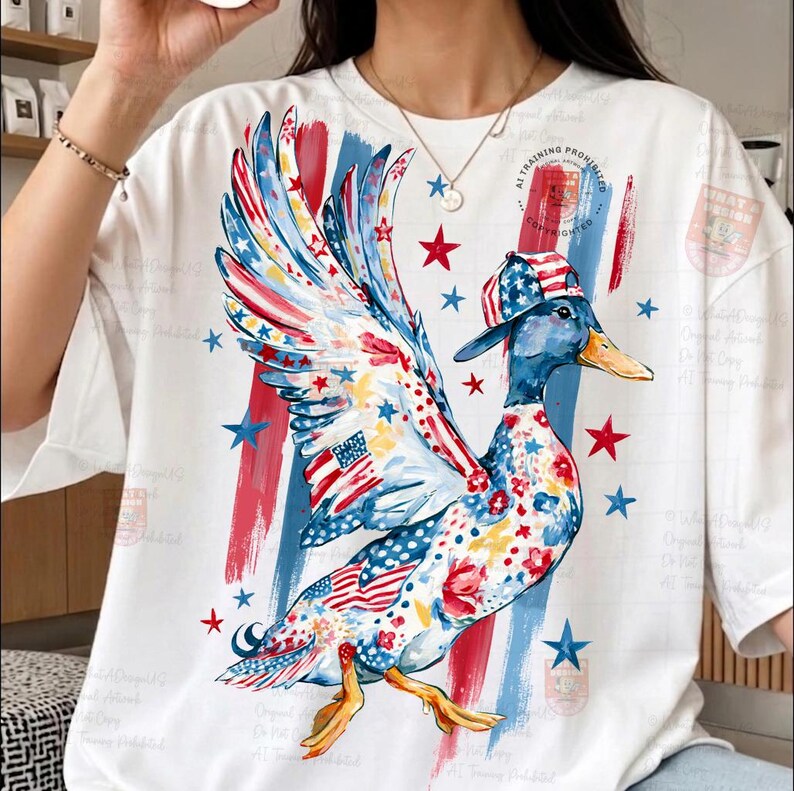 May include: White t-shirt with a quirky duck illustration. The duck is decorated with patriotic motifs, including the American flag and stars, and sports a matching hat. Red and blue brushstrokes and stars complete the design.