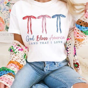 May include: White t-shirt with three red, pink, and blue bows and the text "God Bless America Land That I Love".