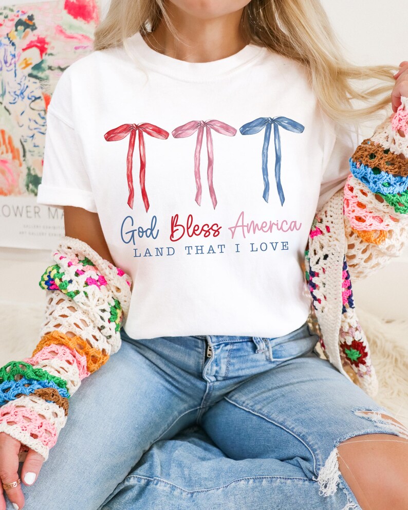 May include: White t-shirt with three red, pink, and blue bows and the text "God Bless America Land That I Love".