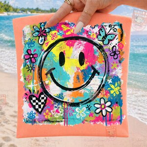 May include: A peach-colored t-shirt featuring a large, colorful smiley face design. The design includes a black smiley face, flowers, a butterfly, and a checkered heart, all in vibrant colors. The shirt is held up by a hand.