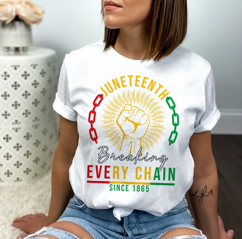 Peut inclure: T-shirt blanc avec un motif graphique repr&eacute;sentant un poing lev&eacute;, des cha&icirc;nes et le texte "Juneteenth Breaking Every Chain Since 1865".