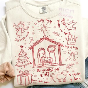 K&ouml;nnte beinhalten: Cremefarbenes Sweatshirt mit rotem Weihnachtsdesign. Das Design zeigt einen Engel, einen Weihnachtsbaum, eine Krippe, ein Geschenk, ein Schaf und die S&auml;tze "Joy to the World", "Holy Night", "Peace on Earth", "King of Kings", "The Greatest Gift of All", "Love Came Down" und "A Savior is Born".