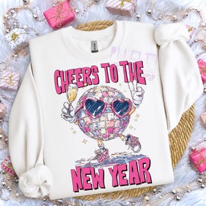 May include: White sweatshirt with a graphic of a disco ball character wearing heart-shaped sunglasses, holding a champagne glass and making a peace sign. The text "CHEERS TO THE NEW YEAR" is printed above and below the disco ball.