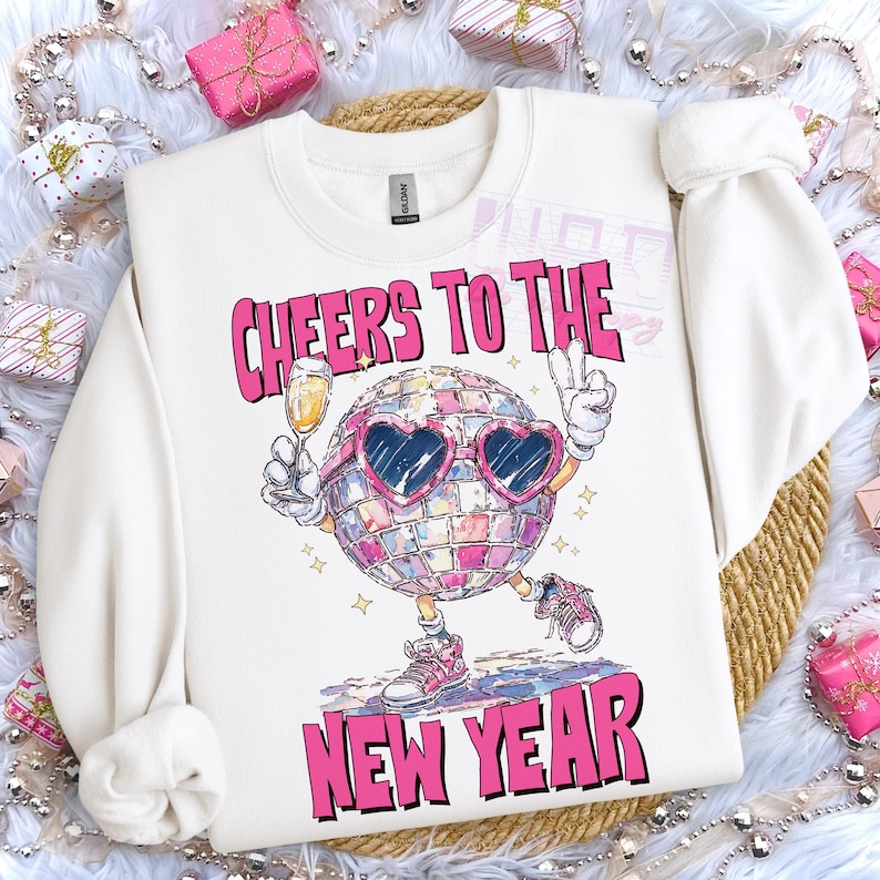 May include: White sweatshirt with a graphic of a disco ball character wearing heart-shaped sunglasses, holding a champagne glass and making a peace sign. The text "CHEERS TO THE NEW YEAR" is printed above and below the disco ball.