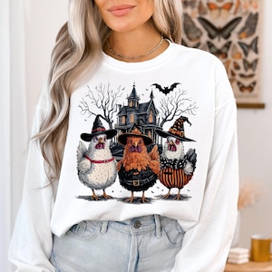 K&ouml;nnte beinhalten: Wei&szlig;er Langarm-Sweatshirt mit einem Grafikdruck von drei H&uuml;hnern, die Hexenh&uuml;te vor einem Spukhaus tragen. Die H&uuml;hner sind wei&szlig;, braun und schwarz-wei&szlig; gestreift. Die Grafik enth&auml;lt auch eine Fledermaus, die &uuml;ber dem Haus fliegt.