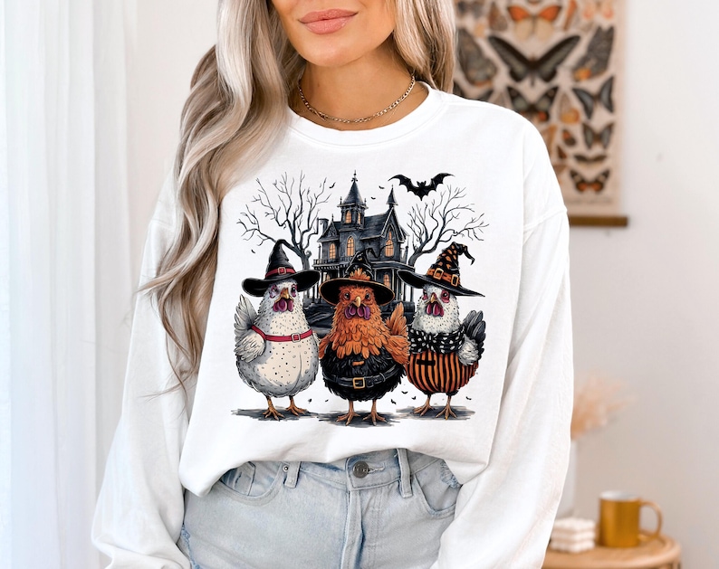 K&ouml;nnte beinhalten: Wei&szlig;er Langarm-Sweatshirt mit einem Grafikdruck von drei H&uuml;hnern, die Hexenh&uuml;te vor einem Spukhaus tragen. Die H&uuml;hner sind wei&szlig;, braun und schwarz-wei&szlig; gestreift. Die Grafik enth&auml;lt auch eine Fledermaus, die &uuml;ber dem Haus fliegt.
