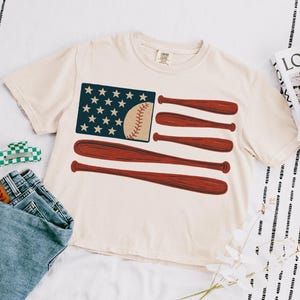 May include: Cream-colored t-shirt featuring a baseball-themed American flag design. The flag graphic includes a blue field with white stars, a baseball, and red baseball bats. The shirt is paired with blue jeans and a green and white hair clip.