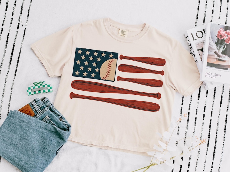 May include: Cream-colored t-shirt featuring a baseball-themed American flag design. The flag graphic includes a blue field with white stars, a baseball, and red baseball bats. The shirt is paired with blue jeans and a green and white hair clip.