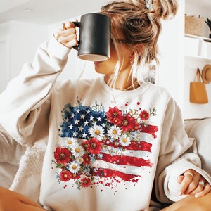 May include: Cream-colored sweatshirt featuring a watercolor design of the American flag adorned with red, white, and blue flowers. The flag design includes stars, stripes, and floral accents, creating a patriotic and artistic look.