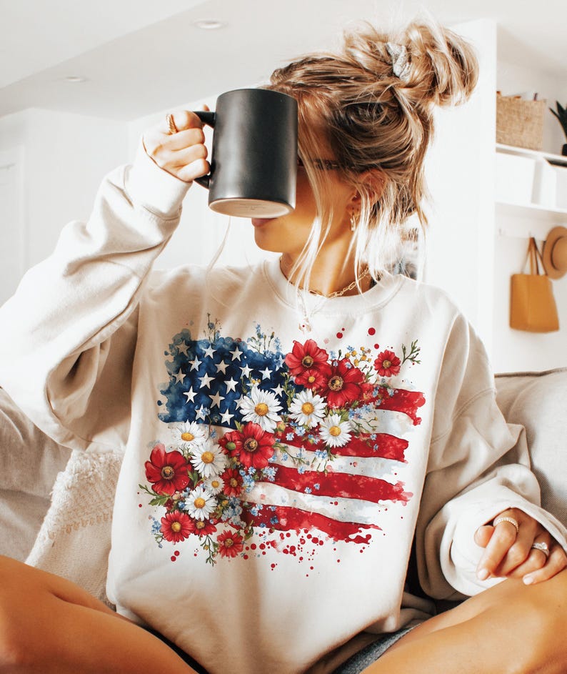 May include: Cream-colored sweatshirt featuring a watercolor design of the American flag adorned with red, white, and blue flowers. The flag design includes stars, stripes, and floral accents, creating a patriotic and artistic look.