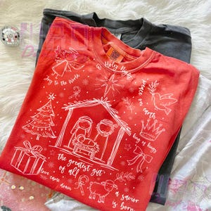 K&ouml;nnte beinhalten: Ein rotes Comfort Colors Crew-Neck-T-Shirt mit einer wei&szlig;en Weihnachtsgrafik. Das Design umfasst eine Krippenszene, einen Engel, einen Weihnachtsbaum und den Text "Joy to the World", "Holy Night" und "The greatest gift of all."