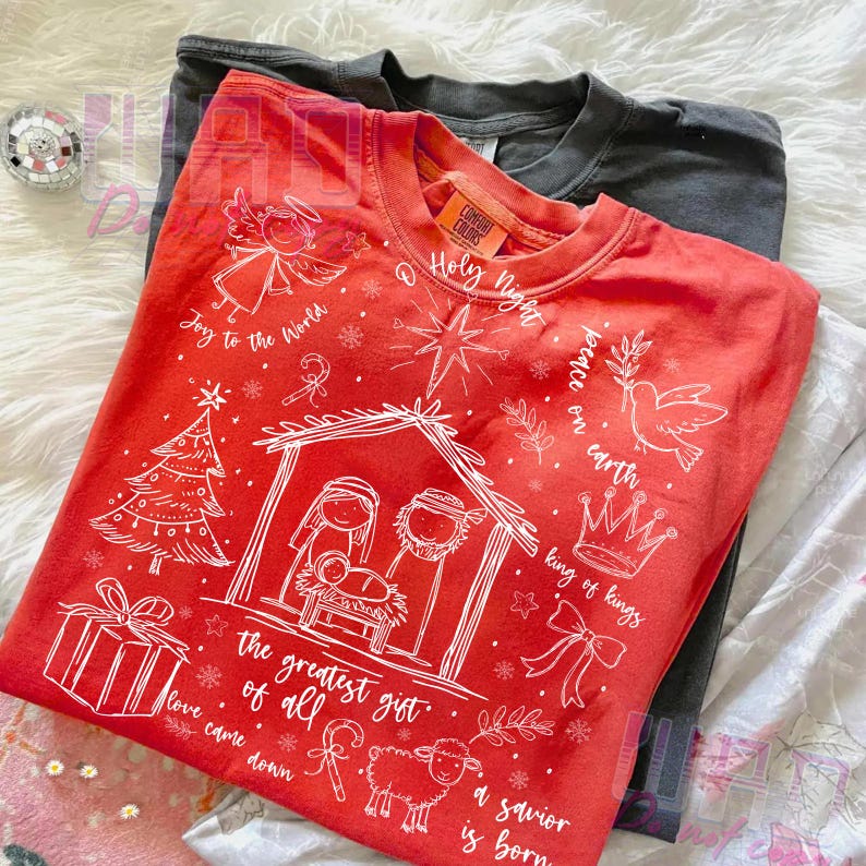 K&ouml;nnte beinhalten: Ein rotes Comfort Colors Crew-Neck-T-Shirt mit einer wei&szlig;en Weihnachtsgrafik. Das Design umfasst eine Krippenszene, einen Engel, einen Weihnachtsbaum und den Text "Joy to the World", "Holy Night" und "The greatest gift of all."