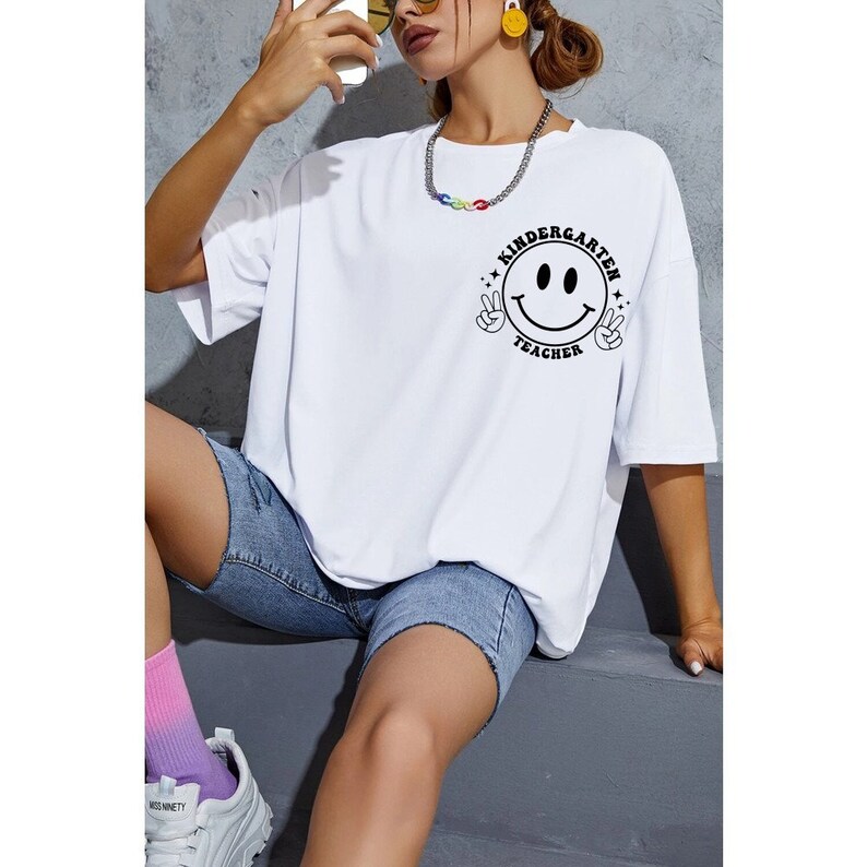 May include: White oversized t-shirt featuring a black smiley face graphic, peace signs, and the text "Kindergarten Teacher". The shirt is worn with denim shorts. The model is wearing sunglasses and a necklace.