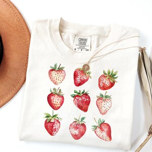 Op de afbeelding: Een wit T-shirt met een afbeelding van negen rode aardbeien. Het shirt heeft een label met de tekst "Comfort Colors" en de maten "S - P - CH".