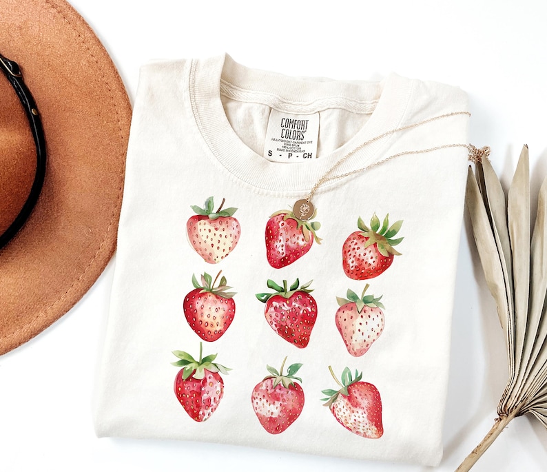 Op de afbeelding: Een wit T-shirt met een afbeelding van negen rode aardbeien. Het shirt heeft een label met de tekst "Comfort Colors" en de maten "S - P - CH".