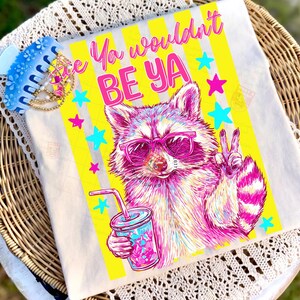 Peut inclure: Un t-shirt blanc avec un motif color&eacute; d'un raton laveur portant des lunettes de soleil roses et tenant une boisson. Le t-shirt porte l'inscription "Ya wouldn't BE YA" avec des rayures jaunes et des &eacute;toiles. Une pince &agrave; cheveux bleue est visible.