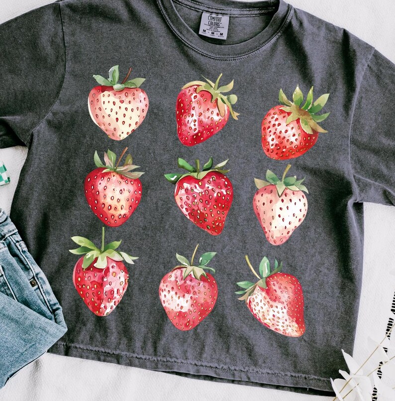 Op de afbeelding: Een donkergrijs T-shirt met een aquarelprint van negen rode aardbeien. De aardbeien zijn in een rasterpatroon op het shirt gerangschikt.