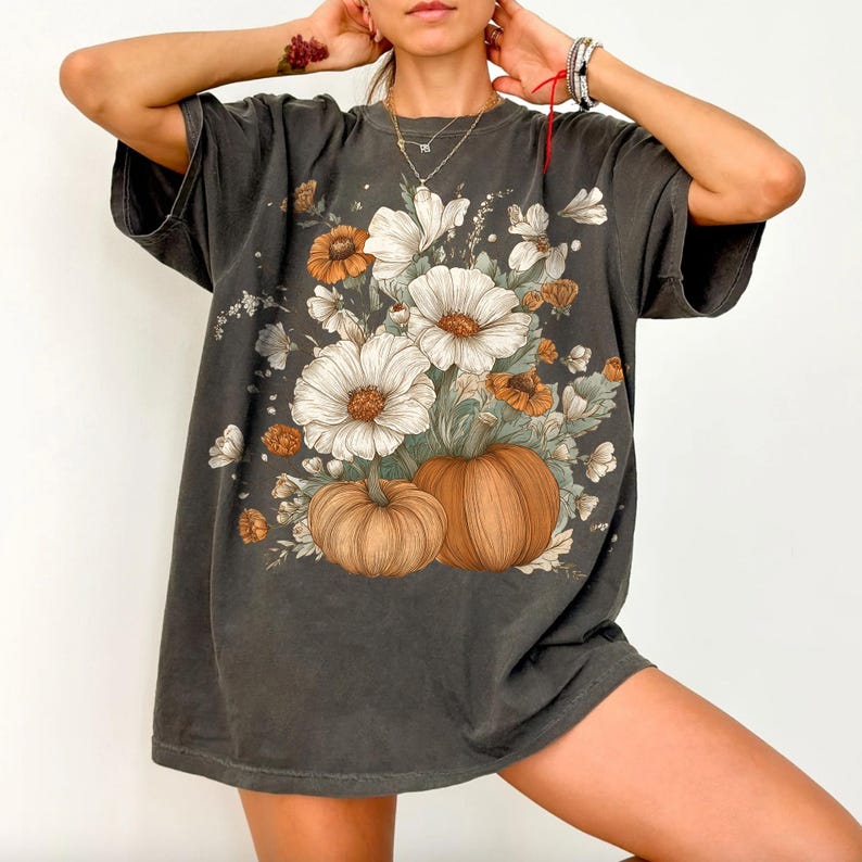 Pu&ograve; includere: T-shirt grigio scuro con grafica floreale e zucca. Il design include grandi fiori bianchi con centri arancioni, fiori arancioni pi&ugrave; piccoli e due zucche marroni. La maglietta ha maniche corte e una vestibilit&agrave; ampia.