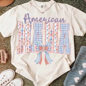 Op de afbeelding: Cr&egrave;mekleurig T-shirt met de woorden "American MAMA" in blauwe letters. De letters zijn versierd met rode en blauwe patronen en een strik. Een zilveren ketting is zichtbaar.