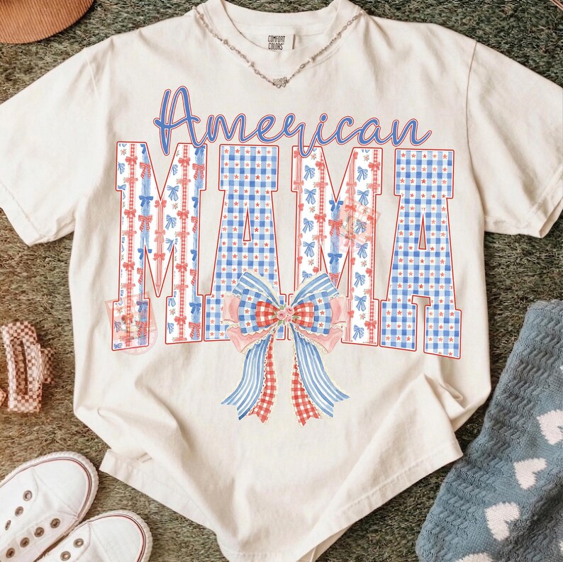 Op de afbeelding: Cr&egrave;mekleurig T-shirt met de woorden "American MAMA" in blauwe letters. De letters zijn versierd met rode en blauwe patronen en een strik. Een zilveren ketting is zichtbaar.