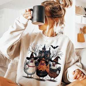 K&ouml;nnte beinhalten: Ein wei&szlig;er Pullover mit einem grafischen Druck von drei H&uuml;hnern, die Hexenh&uuml;te tragen, vor einem Spukhaus. Die H&uuml;hner tragen schwarz, orange und wei&szlig; gestreifte Kleidung.