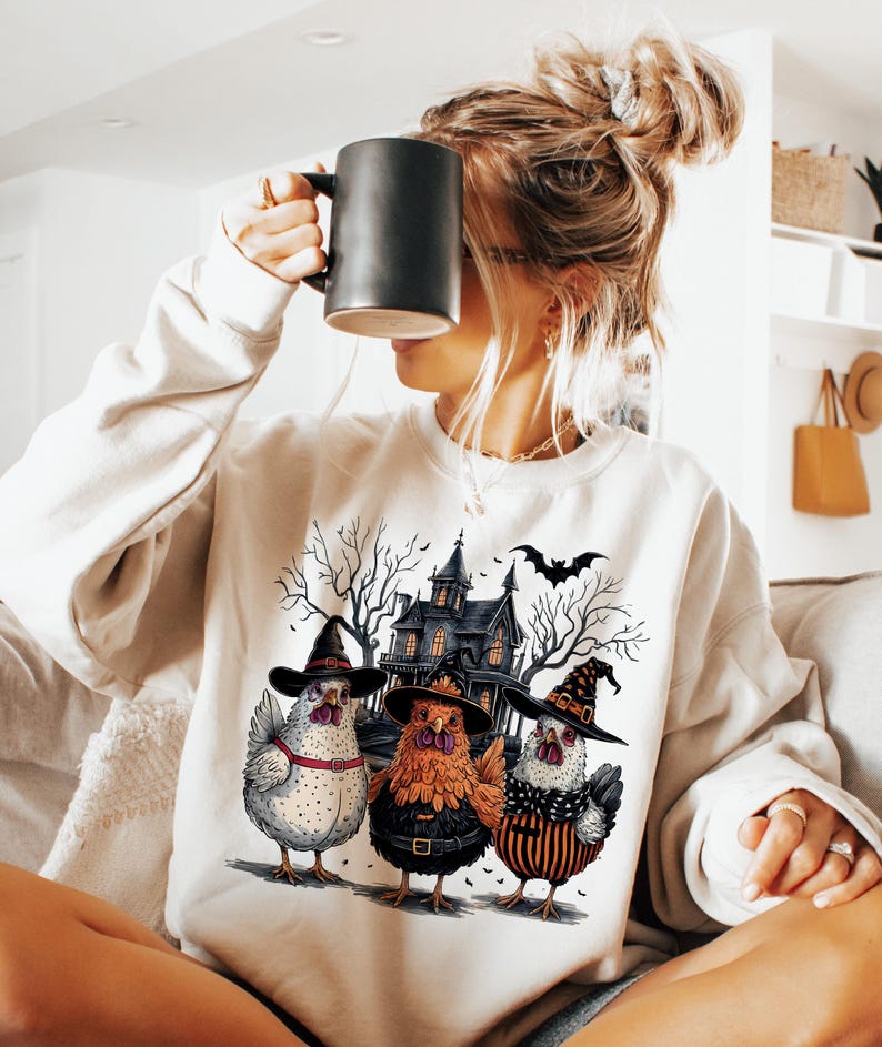 K&ouml;nnte beinhalten: Ein wei&szlig;er Pullover mit einem grafischen Druck von drei H&uuml;hnern, die Hexenh&uuml;te tragen, vor einem Spukhaus. Die H&uuml;hner tragen schwarz, orange und wei&szlig; gestreifte Kleidung.
