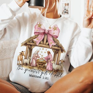 May include: White sweatshirt with a pink bow and a nativity scene graphic. The text "The greatest gift of all" and "Matthew 1:21" are printed below the image.