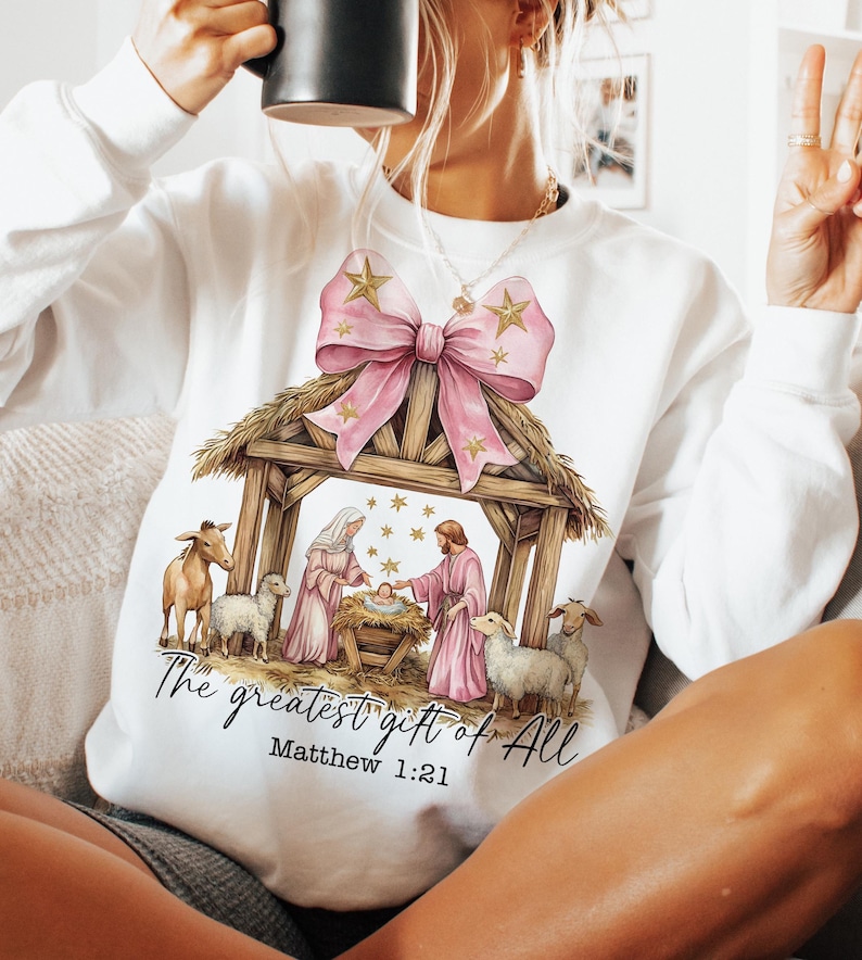 May include: White sweatshirt with a pink bow and a nativity scene graphic. The text "The greatest gift of all" and "Matthew 1:21" are printed below the image.