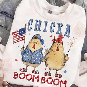 May include: White sweatshirt with a graphic of two cartoon chickens. One chicken holds an American flag, the other a sparkler. The text "CHICKA BOOM BOOM" is printed in blue and red.