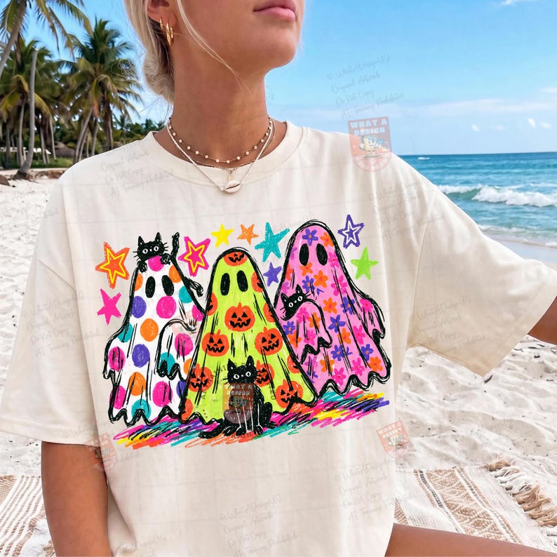 K&ouml;nnte beinhalten: Ein cremefarbenes T-Shirt mit einer Grafik von drei Geistern. Der linke Geist hat lila und orangefarbene Punkte, der mittlere Geist ist limettengr&uuml;n mit orangefarbenen K&uuml;rbissen und der rechte Geist ist rosa mit orangefarbenen Blumen. Schwarze Katzen.