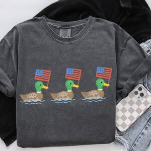 May include: Dark gray t-shirt featuring three ducks, each holding a small American flag. The ducks are depicted swimming on water. The shirt is paired with jeans and a phone with a checkered case.