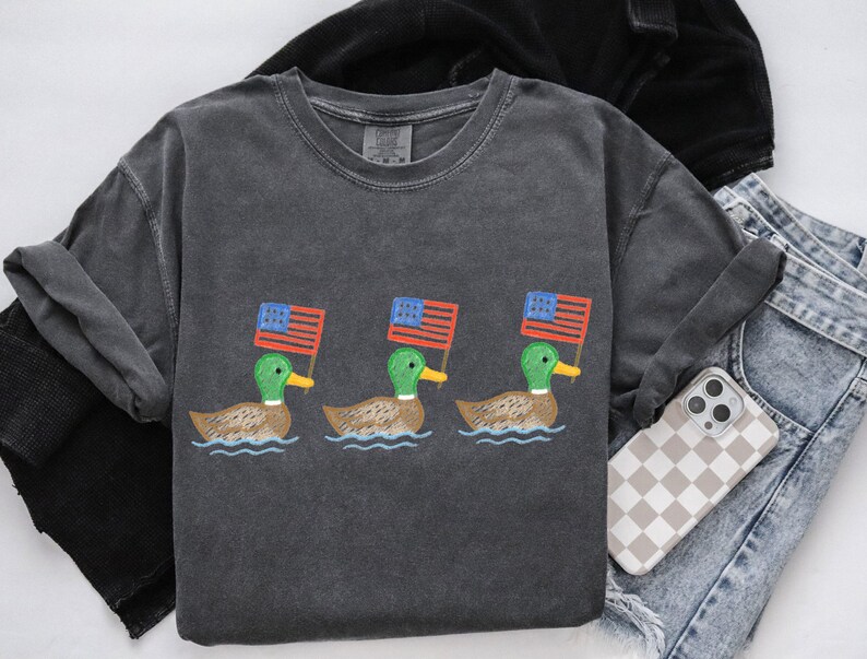 May include: Dark gray t-shirt featuring three ducks, each holding a small American flag. The ducks are depicted swimming on water. The shirt is paired with jeans and a phone with a checkered case.