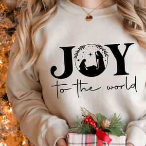 Pu&ograve; includere: Una felpa bianca con le parole "JOY to the world" stampate in nero. La parola "JOY" &egrave; stilizzata con un cerchio che contiene una silhouette nera della scena della Nativit&agrave;.